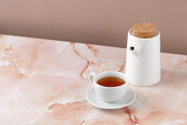Kubek lub filiżanka czarnej herbaty i biały ceramiczny czajniczek na beżowo różowym marmurowym tle Eko minimalizm