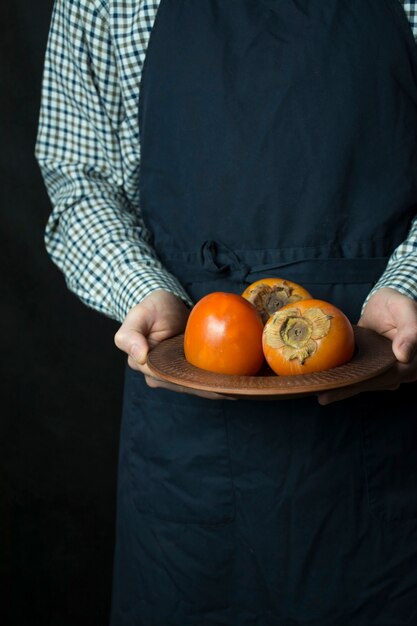 Kucharz trzyma w rękach persimmon. Męskie ręce trzymają talerz ze świeżą persimmon. Owoce sezonowe. Ciemne tło.