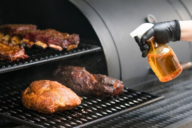 Kuchnia restauracyjna z grillem. Przycięte zdjęcie szefa kuchni opryskującego grillowaną pierś z indyka, wołowinę, mięso wieprzowe i żeberka w palaczu BBQ.