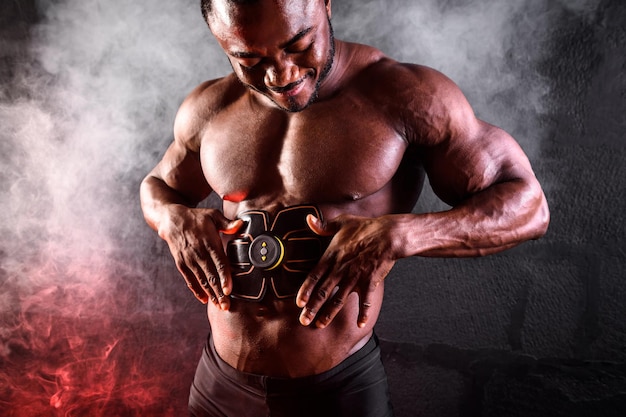 Kulturysta mężczyzna Afrykanin używa elektronicznego pasa stymulatora mięśni trenera mięśni brzucha na czarnym tle z dymem