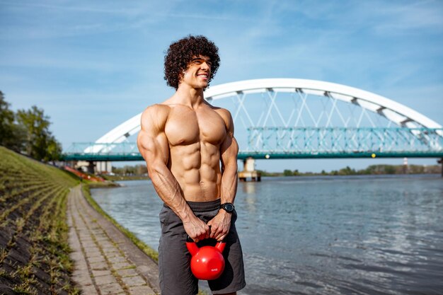 Kulturysta z sześciopakiem, doskonałym ABS, ramionami, bicepsami, tricepsami i klatką piersiową wykonuje mocny trening ciała z odważnikiem kettlebell, w pobliżu rzeki.