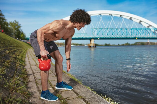 Kulturysta z sześciopakiem, doskonałym ABS, ramionami, bicepsami, tricepsami i klatką piersiową wykonuje mocny trening ciała z odważnikiem kettlebell, w pobliżu rzeki.