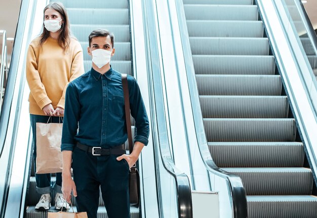 Kupujący w maskach ochronnych stojący na schodach ruchomych w centrum handlowym