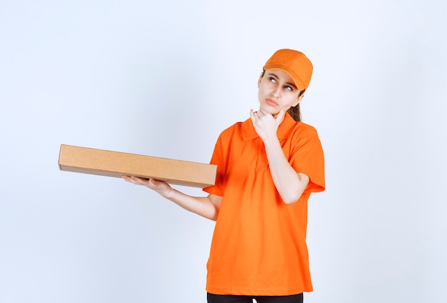 Kurierka w pomarańczowym mundurze trzymająca pudełko z pizzą na wynos i wygląda na zamyśloną lub mającą pomysł