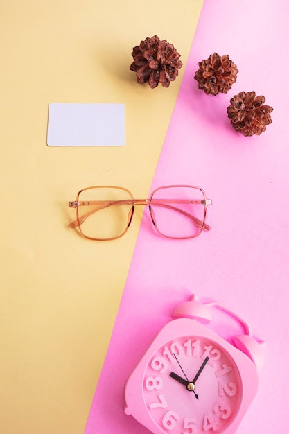 Kwadratowe okulary na zdjęciu w minimalistycznym letnim stylu na pastelowym różowym i żółtym tle. Budzik, kwiaty sosny, wizytówki