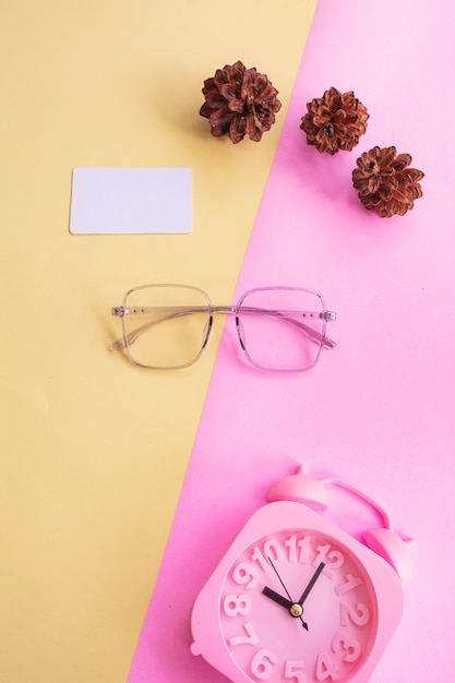 Kwadratowe okulary na zdjęciu w minimalistycznym letnim stylu na pastelowym różowym i żółtym tle. Budzik, kwiaty sosny, wizytówki