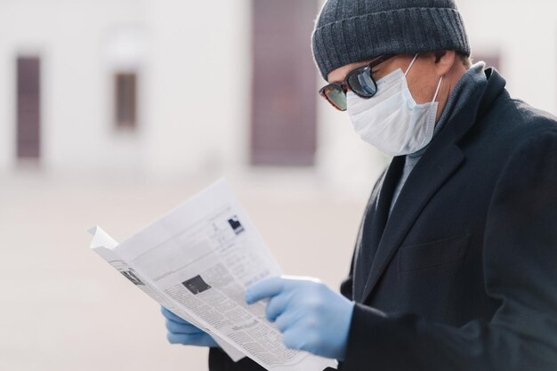 Kwarantanna na koronawirusa Poziome ujęcie dorosłego mężczyzny nosi maskę ochronną i jednorazowe rękawiczki czyta świeżą gazetę dowiaduje się o sytuacji pandemicznej Koncepcja zapobiegania zakażeniom