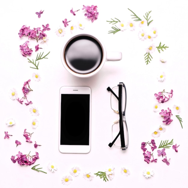 Kwiatki bzu i stokrotka z telefonem komórkowym, okularami i filiżanką kawy na tle biały stół. Płaski układanie, widok z góry