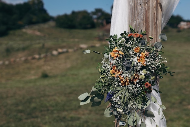 Kwiatowa dekoracja dekoracyjna białego drewnianego łuku ślubnego ze świeżymi kwiatami Gospodarstwo wiejskie boho ceremonia ślubna na świeżym powietrzu Letnia impreza plenerowa Błękitne niebo zielony trawnik Niewyraźne tło