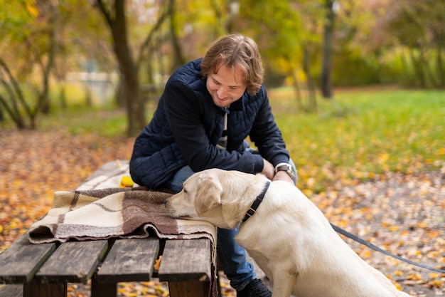 Labrador Wącha Drewnianą ławkę W Parku. Mężczyzna Właściciel Siedzi W Parku Z Białym Retriverem. Zbliżenie.