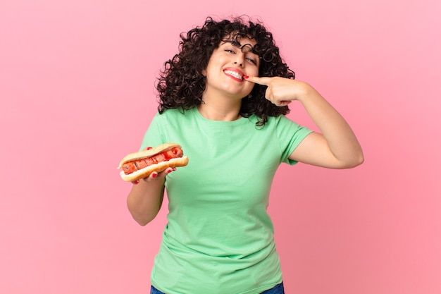 Ładna arabska kobieta, uśmiechnięta pewnie, wskazując na swój szeroki uśmiech i trzymająca hot doga