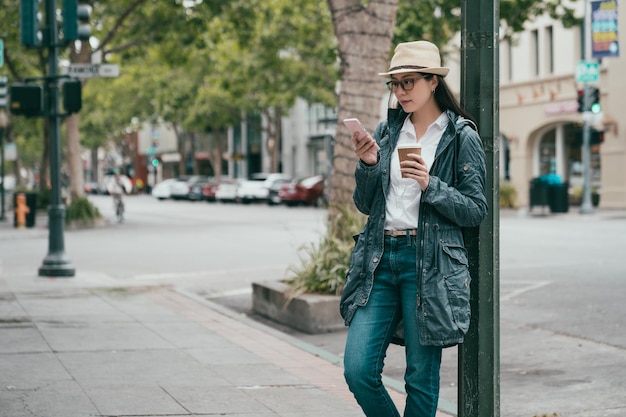 ładna azjatycka kobieta, szukając na telefonie i opierając się na świetle dziennym, relaksując się na ulicy.