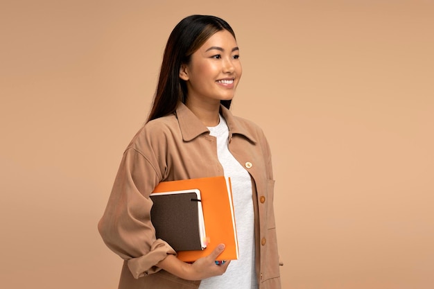 Ładna azjatycka studentka trzymająca książki lub papierowe organizery odwracająca wzrok z uśmiechem