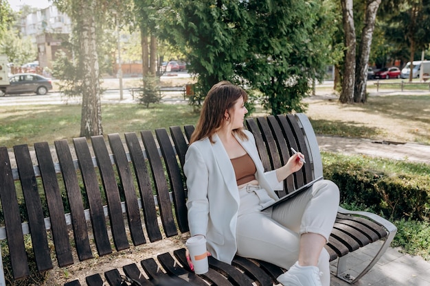 Ładna Biznesowa Kobieta W Białym Garniturze Siedzi W Miejskim Parku Pije Kawę I Pracuje Na Cyfrowym Tablecie