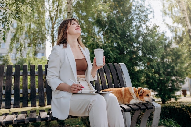 Ładna Bizneswoman Grafik W Białym Garniturze Siedzi W Parku Miejskim Pije Kawę I Pracuje Na Cyfrowym Tablecie Z Psem Welsh Corgi Pembroke