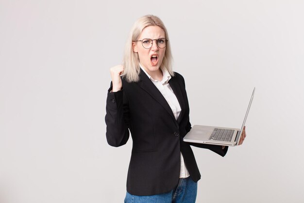 Ładna blond kobieta krzyczy agresywnie ze złym wyrazem twarzy i trzyma laptopa