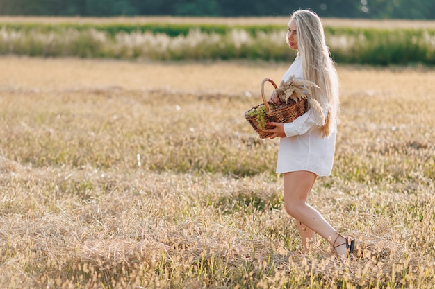 Ładna Blond Kobieta Z Długimi Włosami Na Polu O Zachodzie Słońca Niesie Kosz Owoców I Bukiet Słomy. Lato, Rolnictwo, Przyroda I świeże Powietrze Na Wsi.