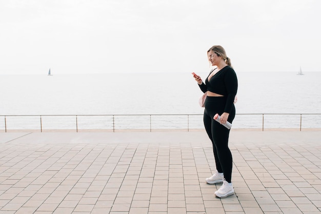 Ładna blondynka plus size odpowiadająca na wiadomości lub patrząca na swój telefon komórkowy przed plażą ubrana w sportowe ubrania