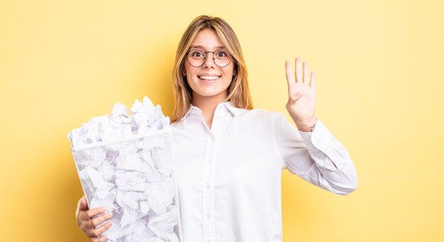 Zdjęcie Ładna blondynka uśmiecha się i wygląda przyjaźnie, pokazując numer cztery. koncepcja śmieci z kulkami papierowymi