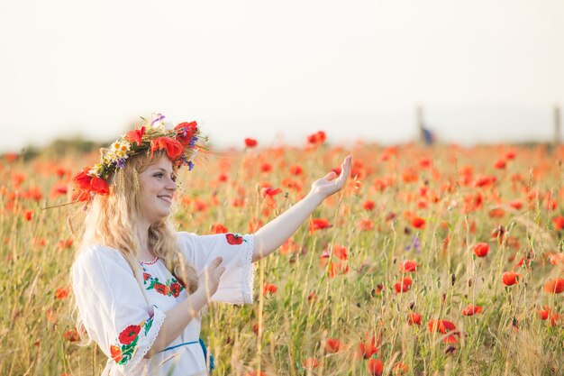 Ładna blondynka w sukience z wzorem maku iz wieńcem na głowie ciesząc się polem z makami