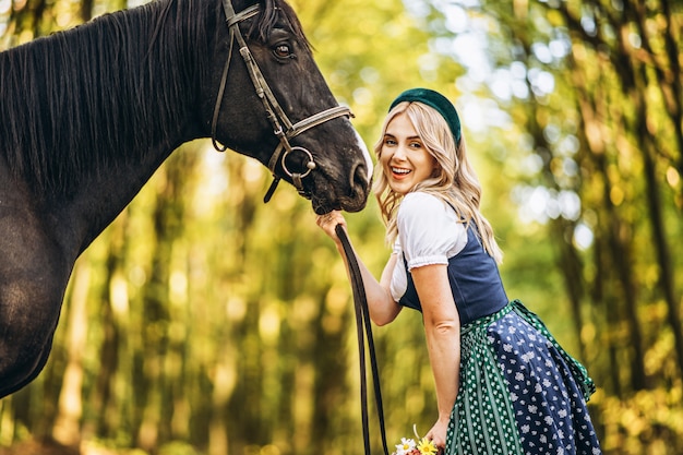 Ładna blondynka w tradycyjnym stroju chodzenie z dużym czarnym koniem w lesie