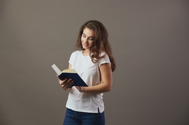 Ładna brązowowłosa dziewczyna ubrana w białą koszulkę i dżinsy czyta książkę na szarym tle.