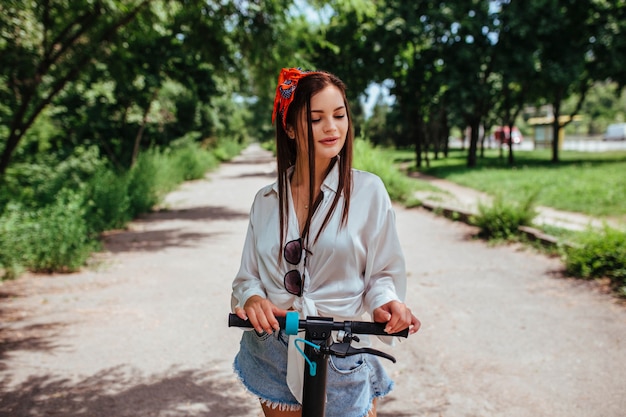 Ładna brunetka dziewczyna jeździ na hulajnodze elektrodowej w parku, w której ma na sobie białą koszulę. koncepcja ekologicznego transportu i wynajmu.