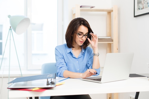 Ładna brunetka dziewczyna w niebieskiej koszuli siedzi przy stole w biurze. Pisze na laptopie i trzyma czarne okulary.