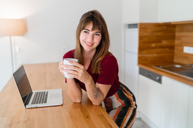 Ładna brunetka kobieta pije kawę i siedzi w nowoczesnej kuchni i korzysta z laptopa. Szukam pracy online