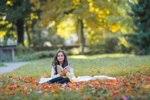 Ładna ciemnowłosa 10-letnia dziewczynka w dzianinowym swetrze czyta książkę siedząc na polanie w jesiennym parku Piknik w przyrodzie Szczęśliwe dziecko się uczy