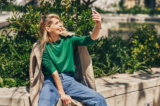 Ładna dziewczyna co autoportret z zabawnym grymasem Młoda radosna kobieta uśmiechając się szeroko trzymając smartfon spacerując na świeżym powietrzu w mieście w słoneczny dzień Szczęśliwa kobieta biorąc selfie na telefon komórkowy