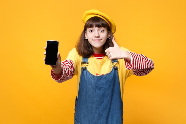 Ładna dziewczyna nastolatek w francuskim berecie pokazując kciuk do góry, trzymając telefon komórkowy z pustym pustym ekranem na białym tle na tle żółtej ściany. Ludzie szczere emocje, koncepcja stylu życia. Makieta miejsca na kopię.
