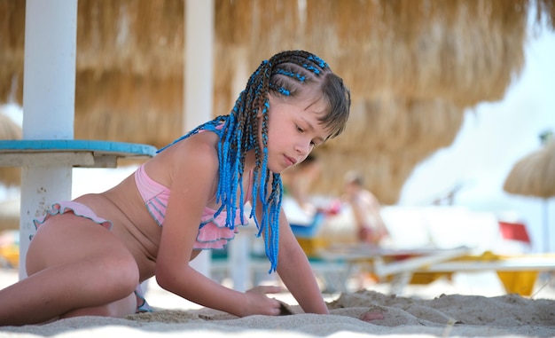 Zdjęcie Ładna dziewczyna preteen w stroju kąpielowym bikini bawiąca się piaskiem w cieniu parasola podczas letnich wakacji tropikalnych