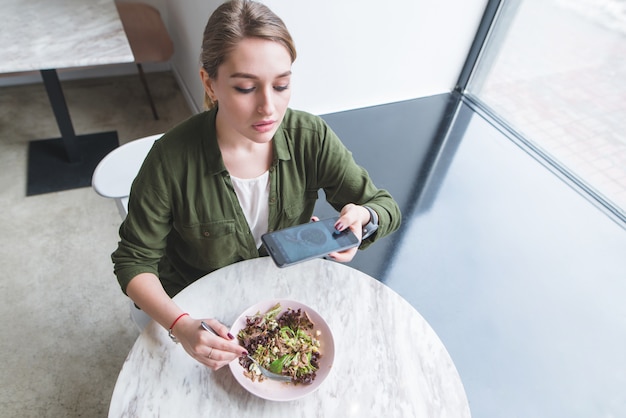 Zdjęcie Ładna dziewczyna robi zdjęcie sałatki w restauracji. blogger odbiera jedzenie na stole w pobliżu okna