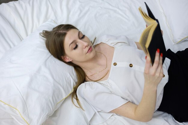 ładna dziewczyna trzyma książkę w dłoniach i czyta leżąc w białym łóżku, czytając i relaksując się, koncepcja wolnego czasu i relaksu w domu