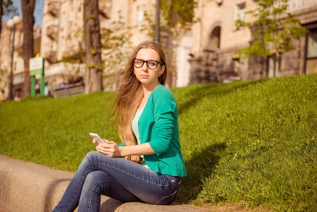 Ładna dziewczyna w okularach siedzi w parku i trzymając smartfon