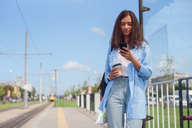 Ładna dziewczyna z filiżanką kawy czeka rano na autobus lub tramwaj na stacji transportu publicznego. Młoda kobieta z inteligentnego telefonu monitorowania transportu za pośrednictwem aplikacji.