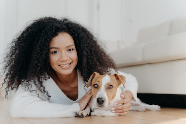 Ładna dziewczyna z włosami afro leży na podłodze z psem wyraża przyjemne emocje pozach w salonie przy kanapie kupił zwierzaka w nowym mieszkaniu Gospodyni z ukochanym zwierzakiem w domu dzieli się dobrą chwilą