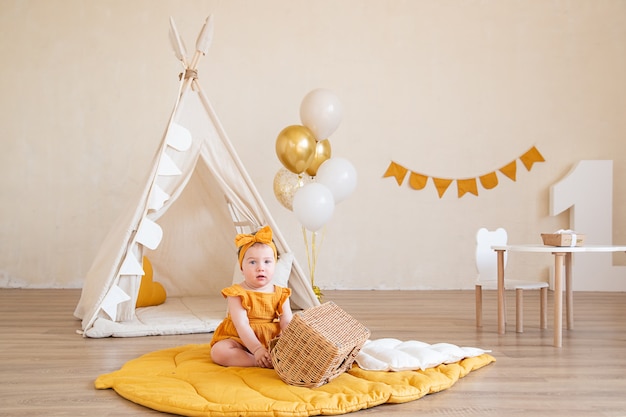Ładna jednoroczna dziewczynka w żółtym ubraniu siedzi na podłodze i bawi się koszem. Studio fotograficzne na urodziny dziecka.