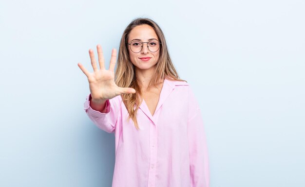 Ładna kaukaska kobieta, uśmiechnięta i wyglądająca przyjaźnie, pokazująca cyfrę piątą lub piątą z ręką do przodu, odliczając w dół