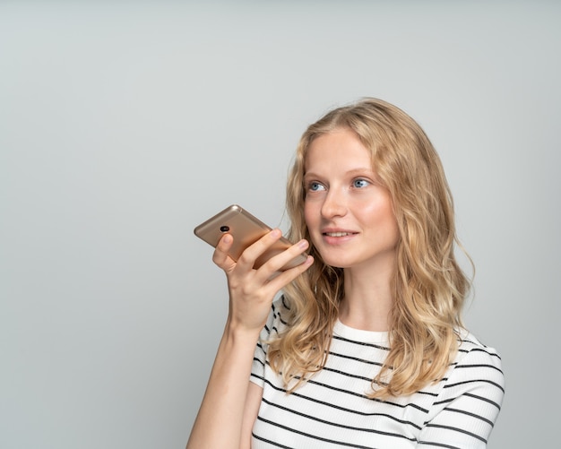 Ładna kobieta dyktuje wiadomość głosową na telefon komórkowy stojący w pobliżu białej ściany, szare tło.