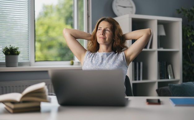 Ładna kobieta freelancer siedzi na wygodnym krześle z rękami za głową i marzy o wakacjach Dojrzała kobieta używa laptopa do pracy w domu