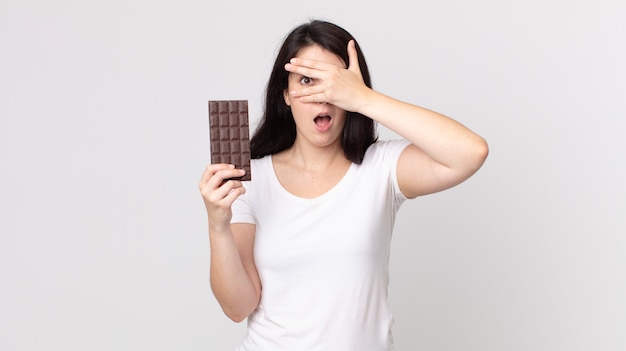 Ładna kobieta, która wygląda na zszokowaną, przestraszoną lub przerażoną, zakrywa twarz dłonią i trzyma tabliczkę czekolady