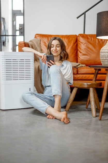 Ładna kobieta siedzi w pobliżu urządzenia oczyszczającego powietrze i nawilżającego w pobliżu kanapy za pomocą smartfona. Pojęcie mikroklimatu zdrowia w domu.