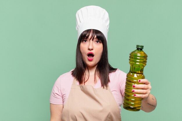 Ładna kobieta szefa kuchni przestraszona wyrazem twarzy i trzymająca butelkę oliwy z oliwek