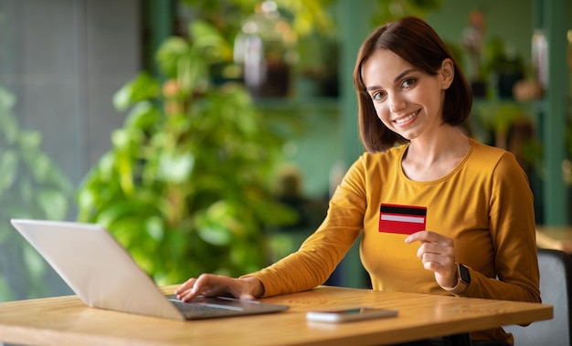 Ładna kobieta trzyma kartę kredytową i używa laptopa w kawiarni