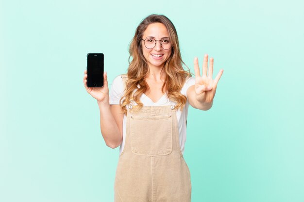 Ładna kobieta uśmiechnięta i wyglądająca przyjaźnie, pokazująca numer cztery i trzymająca smartfona
