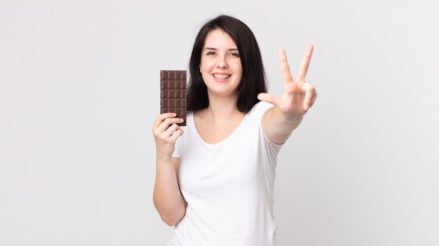 Ładna kobieta uśmiechnięta i wyglądająca przyjaźnie, pokazująca numer trzy i trzymająca tabliczkę czekolady