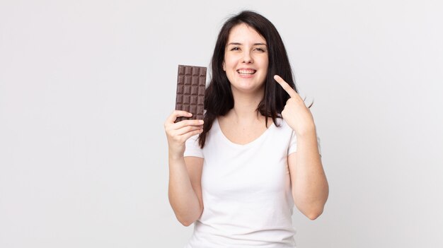 Ładna kobieta, uśmiechnięta pewnie, wskazując na swój szeroki uśmiech i trzymająca tabliczkę czekolady