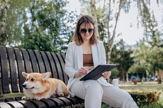 Ładna kobieta w białym garniturze siedzi w parku miejskim z psem Welsh Corgi Pembroke i pracuje na cyfrowym tablecie xA